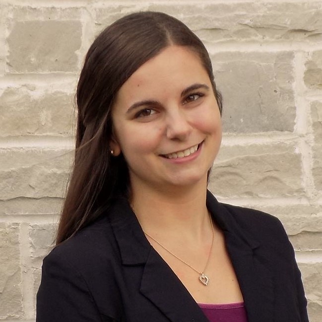 A female staff member stands outside a brick building in a black blazer and maroon shirt, smiling at the camera.